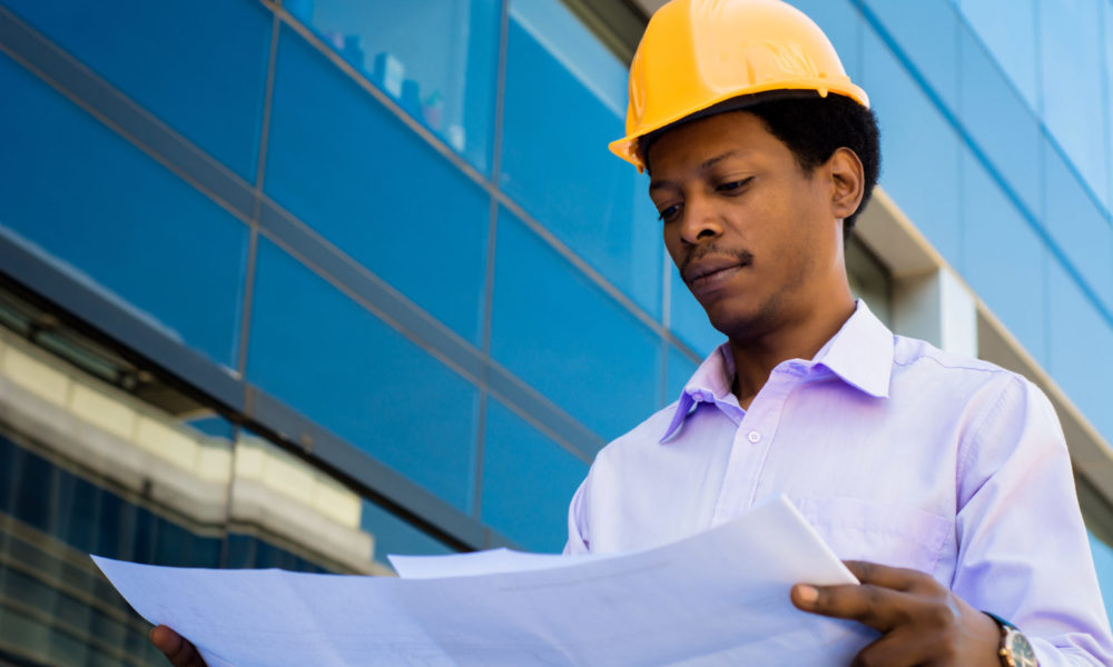 Portrait of professional architect in helmet looking at blue prints outside modern building. Engineer and architect concept.